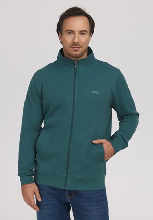 Mężczyzna w turkusowej kurtce na zamek z wysokim kołnierzem i niebieskich dżinsach, stoi z jedną ręką w kieszeni kurtki na jasnym tle.