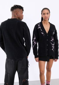 Μαύρο κασκόλ με ροζ σχέδια φλόγας στο μπρο�στινό μέρος. Διαθέτει v-λαιμό, κλείσιμο με κουμπιά και υφή πλεκτού υλικού. Φορεμένο με σκισμένα παντελόνια.