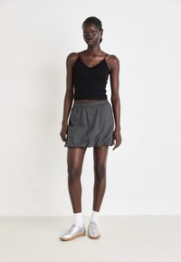 Schwarzes, gestricktes Tanktop mit Spitzenbesatz, kombiniert mit einem grauen karierten Rock. Das Modell trägt silberne Sneakers und weiße Socken. Einfache Hintergrund.