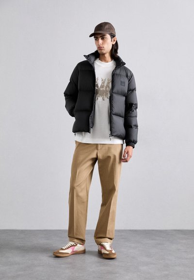 Veste noire rembourrée avec un col haut, t-shirt graphique gris, pantalon beige, et baskets beige avec des accents roses. Design moderne et fonctionnel.