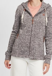 Felpa grigia zipata con texture, cordini bianchi e dettagli in pelle marrone. Indossata sopra una maglietta rosa chiaro e abbinata a pantaloni bianchi.