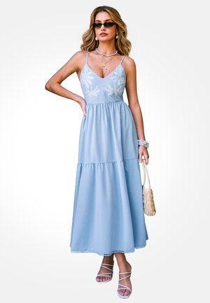 Žena u svijetloplavoj maksi haljini bez rukava s vezom, sunčanim naočalama, slojevitim ogrlicama i bijelim sandalama, držeći pletenu torbu.