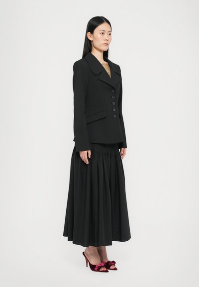 Black tailored blazer with large collar and buttons, paired with a flowing black skirt. Finished with red heeled shoes featuring bows.