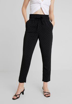 Kobieta w czarnych spodniach o wysokim stanie, zwężanych ku dołowi, z małymi białymi kropkami, z paskiem w talii, białym skręcanym crop topie i czarnych sandałkach na obcasie.