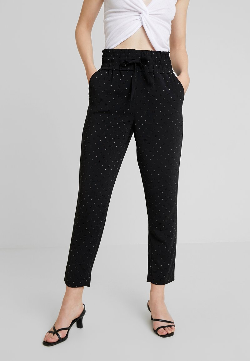 Femme portant un pantalon noir taille haute à coupe fuselée avec de petits pois blancs, une ceinture à nouer, un top court blanc noué et des sandales à talons noires.