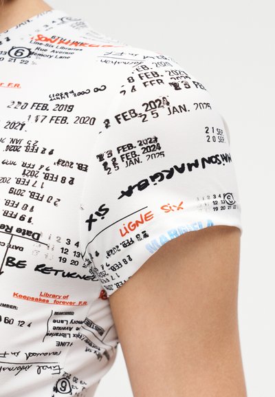 Trumpas baltos spalvos audinio marškinėlis su įvairiais juodais, raudonais ir mėlynais spaudiniais, įskaitant tekstus, datas ir simbolius.