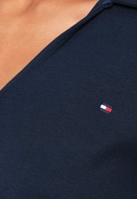 Mørkeblå strikket topp med v-hals, med et lite brodert logo i rødt, hvitt og blått på venstre side nær skulderen.