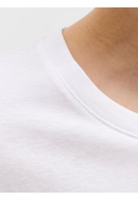 Κοντινή λήψη του λαιμού και του ώμου ενός ατόμου που φοράει απλό λευκό μπλουζάκι με στρογγυλή λαιμόκοψη σε ανοιχτό φόντο.