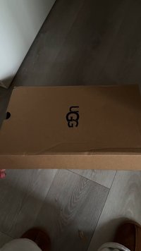 Brown UGG shoe box held by hand above light wood floor, with part of legs and brown slippers visible at the bottom.