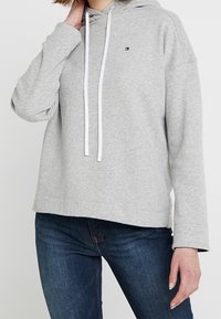Felpa grigia con cappuccio e cordino, dotata di un piccolo logo sul petto e con una vestibilità comoda. Abbinata a jeans blu scuro.