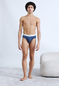 Ανδρικά εσώρουχα σλιπ σε σκούρο μπλε ύφασμα με λευκή ελαστική μέση. Το μοντέλο στέκεται ξυπόλητο σε ανοιχτό γκρι χαλί, κοιτώντας μπροστά.