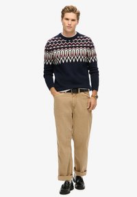 Marineblauer Strickpullover mit einem mehrfarbigen Fair-Isle-Muster, kombiniert mit hellbraunen Hosen und schwarzen Schuhen, die einen Gürtel und aufgerollte Ärmel aufweisen.