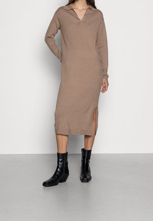 Frau trägt ein braunes, langärmliges Strickmidi-Kleid mit Kragen und seitlichem Schlitz, kombiniert mit schwarzen Stiefeletten.