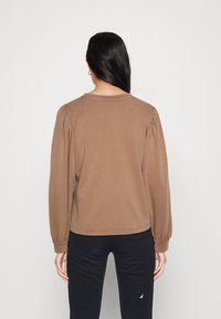 Felpa marrone con maniche lunghe e a sbuffo; scollatura rotonda e vestibilità comoda. Realizzata in un tessuto morbido e strutturato. Pantaloni neri visibili sotto.