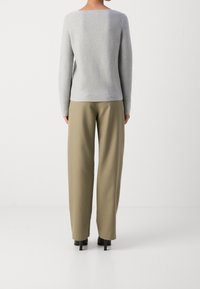 Henkilö seisoo selkä kameraan päin, yllään vaaleanharmaa pitkähihainen paita, oliivinvihreät leveälahkeiset housut ja mustat korokepohjaiset kengät.