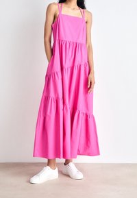 Różowa sukienka maxi z warstwowym krojem, kwadratowym dekoltem i cienkimi ramiączkami, wykonana z lekkiego materiału. Noszona z białymi trampkami.