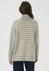 Model wears a gray and cream striped sweater with a high collar, viewed from the back, paired with dark blue jeans against a plain background.