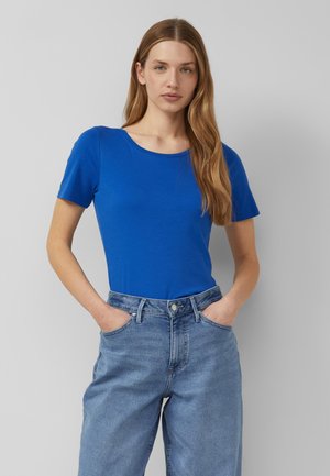 Frau mit langen blonden Haaren, die ein hellblaues kurzärmeliges Shirt und hellblaue High-Waist-Jeans trägt, mit den Händen in den Taschen.