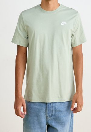 Mann trägt ein hellgrünes Nike-T-Shirt und blaue Jeans, steht vor einem einfarbigen weißen Hintergrund.