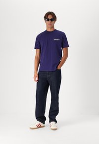 Camiseta de algodón morada con un logo en el pecho, combinada con jeans de color azul oscuro y zapatillas blancas con rayas rojas, llevada con gafas de sol.