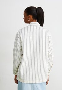 Veste à rayures blanches avec manches longues, col et boutons aux poignets. Présente des lignes verticales foncées et une coupe décontractée.