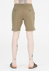 Olivgrön bomullsshorts med uppvikta fållar, med två bakfickor med knappstängningar. Bärs med bruna slipskor.