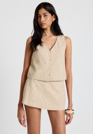 Frau trägt ein beige ärmelloses, geknöpftes Oberteil und einen passenden kurzen Rock mit dezenten weißen Stickmustern, accessoriert mit Armreifen.