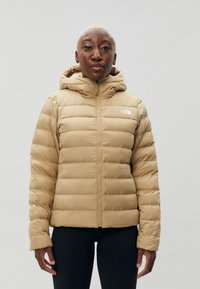 Chaqueta acolchada beige con diseño acolchado, cremallera completa y capucha. Presenta el logo de The North Face en el pecho izquierdo. Tejido suave y ligero.