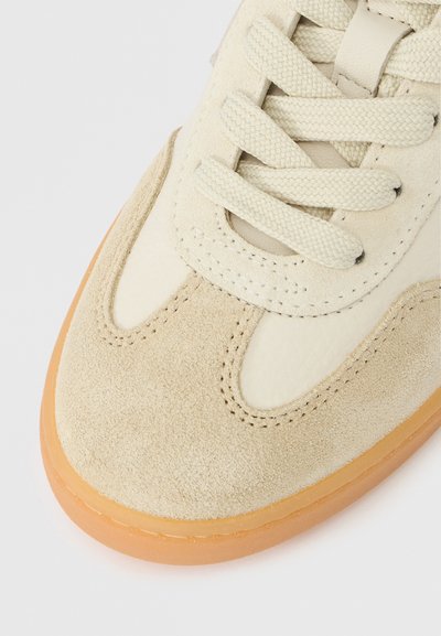 Μπεζ αθλητικό παπούτσι με δερμάτινα και σουέντ υλικά, στρογγυλή μύτη, υφασμένο πάνω μέρος, επίπεδα κορδόνια και σόλα από καουτσούκ σε χρώμα καφέ. Ορατή ραφή στη λεπτομέρεια.