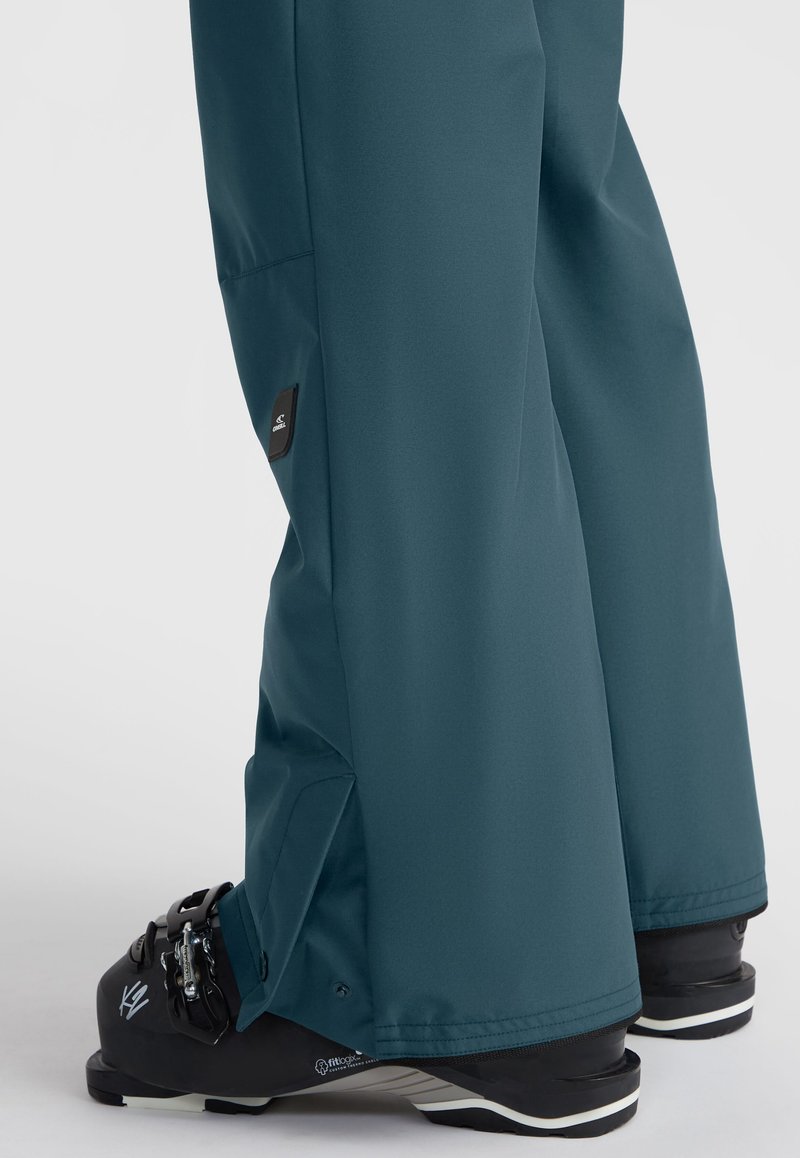 Turquoise skibroek met een gladde textuur, voorzien van een verstelbare pijp en een zwart logo-opdruk aan de zijkant. GStyled met zwarte skischoenen.