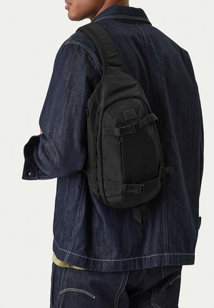 Osoba nosící tmavou džínovou bundu a kalhoty s černým batůžkem na jedno rameno, který má na zádech dva popruhy se sponami.