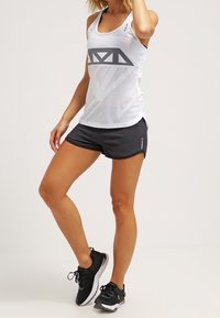 Débardeur de sport blanc avec un grand motif de losange gris, associé à un short texturé noir et des chaussures de course noires.