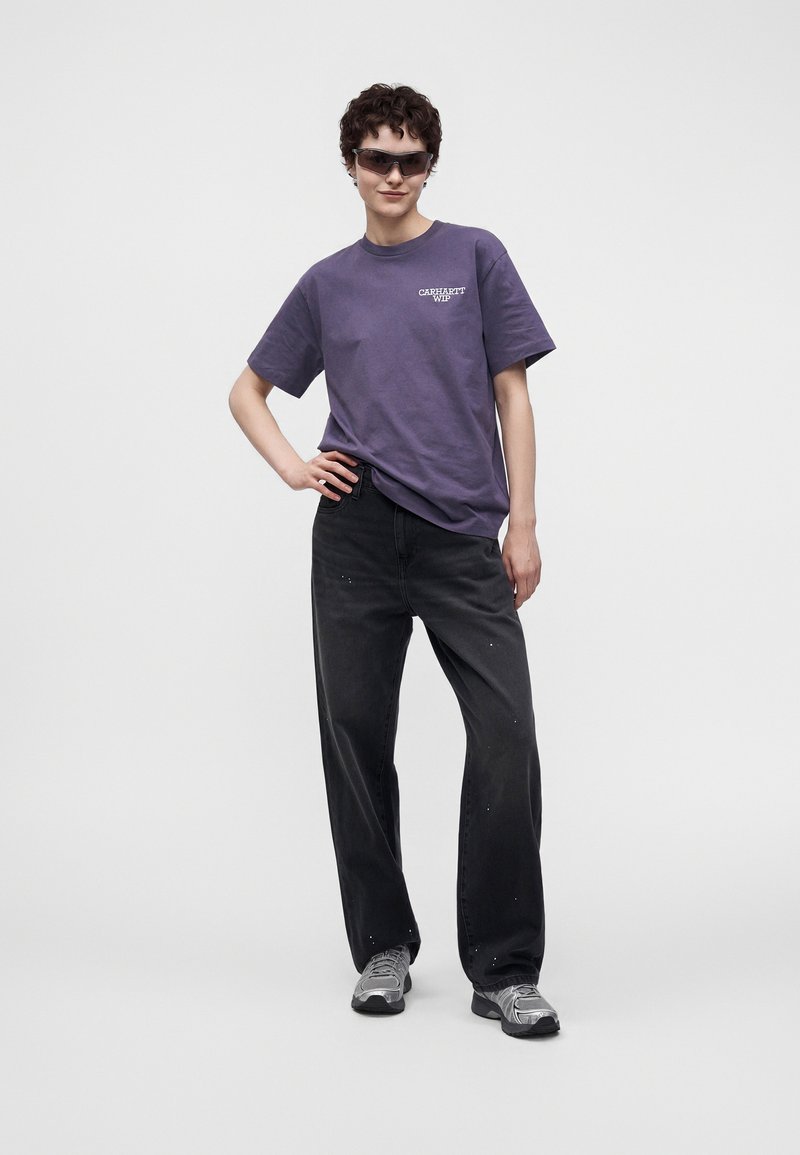 Persona che indossa occhiali da sole scuri, una t-shirt Carhartt viola, jeans neri larghi con macchie di vernice e sneaker grigie, in piedi con una mano sul fianco.