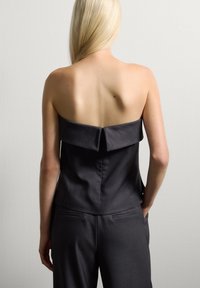 Persona bionda con lunghi capelli, che indossa un top nero senza spalline e pantaloni neri, in piedi con le mani in tasca contro un muro bianco.