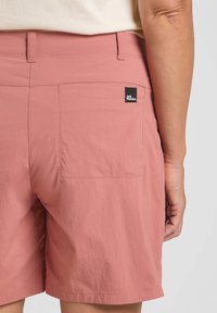 Pantaloni shorts rosa realizzati in tessuto leggero, con una tasca posteriore dotata di un'etichetta logo nera e dettagli di cucitura pulita. Texture morbida e design casual.