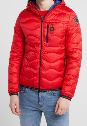 Man wearing a bright red quilted jacket with black zipper and patch emblem on sleeve, paired with light blue jeans.