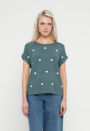 Turkis kortærmet t-shirt med hvide hjerteprint; løs pasform og oprullede ærmer. Kombineret med løse blå denimjeans.