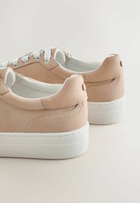 Paire de baskets basses beige avec lacets blancs et semelles épaisses en caoutchouc blanc sur un fond clair.