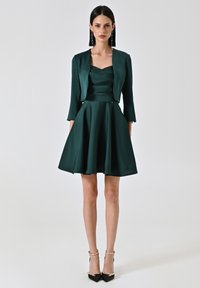 Abito in satin verde smeraldo con corpino aderente e gonna svasata, accompagnato da una giacca corta; presenta maniche a tre quarti e scarpe con tacco appuntito.