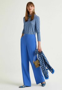 Top blu a maniche lunghe con motivo geometrico, abbinato a pantaloni larghi blu. Dettagli includono una borsa dorata e una sciarpa a motivo.