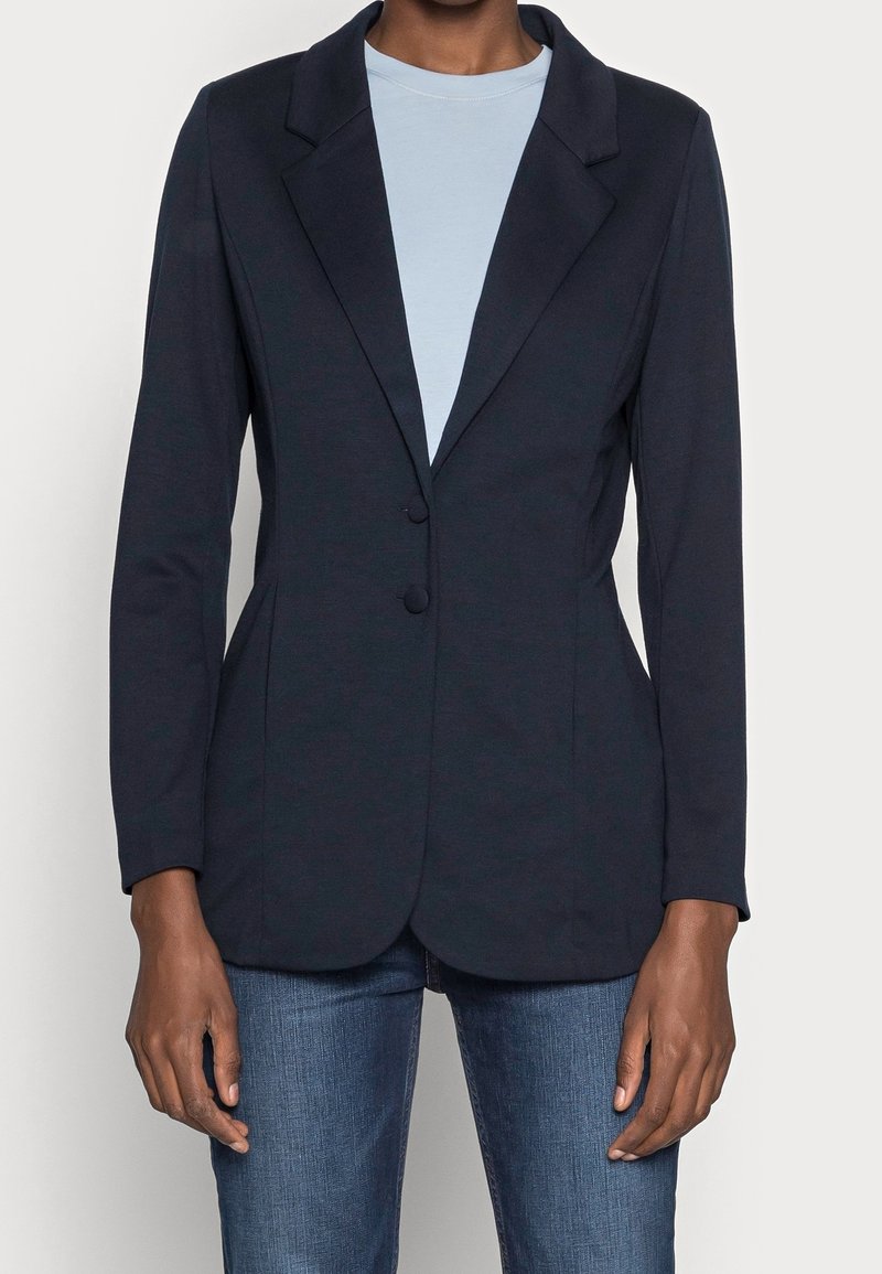 Personne portant un blazer bleu marine ajusté à deux boutons sur une chemise bleu clair et un jean bleu, debout devant un fond uni.