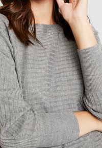 Kobieta w szarym prążkowanym swetrze, z jedną ręką założoną na piersi, a drugą przy twarzy, na jednolitym tle.