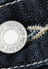 Bouton en métal avec inscription "WEEKDAY" attaché à un tissu en denim bleu foncé, présentant des coutures blanches et une surface texturée.