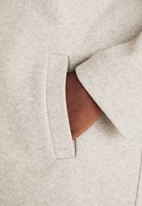 Jasnoszary wełniany płaszcz z boczną kieszenią wyłożoną miękką, brązową podszewką oraz kołnierzem z wycięciem. Tkanina ma teksturowany, filcowy wygląd.