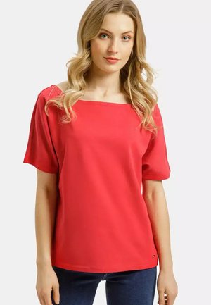 Blondynka o falowanych włosach, ubrana w luźny czerwony top z krótkim rękawem i ciemnoniebieskie dżinsy, stojąca na jednolitym białym tle.