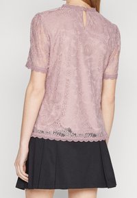 Femme portant un haut en dentelle mauve à manches courtes avec des motifs floraux, associé à une jupe plissée noire, vue de dos.