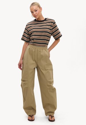 Femme aux cheveux blonds en chignon portant un t-shirt rayé marron, un pantalon beige ample et des sandales foncées, debout avec une main sur la hanche.