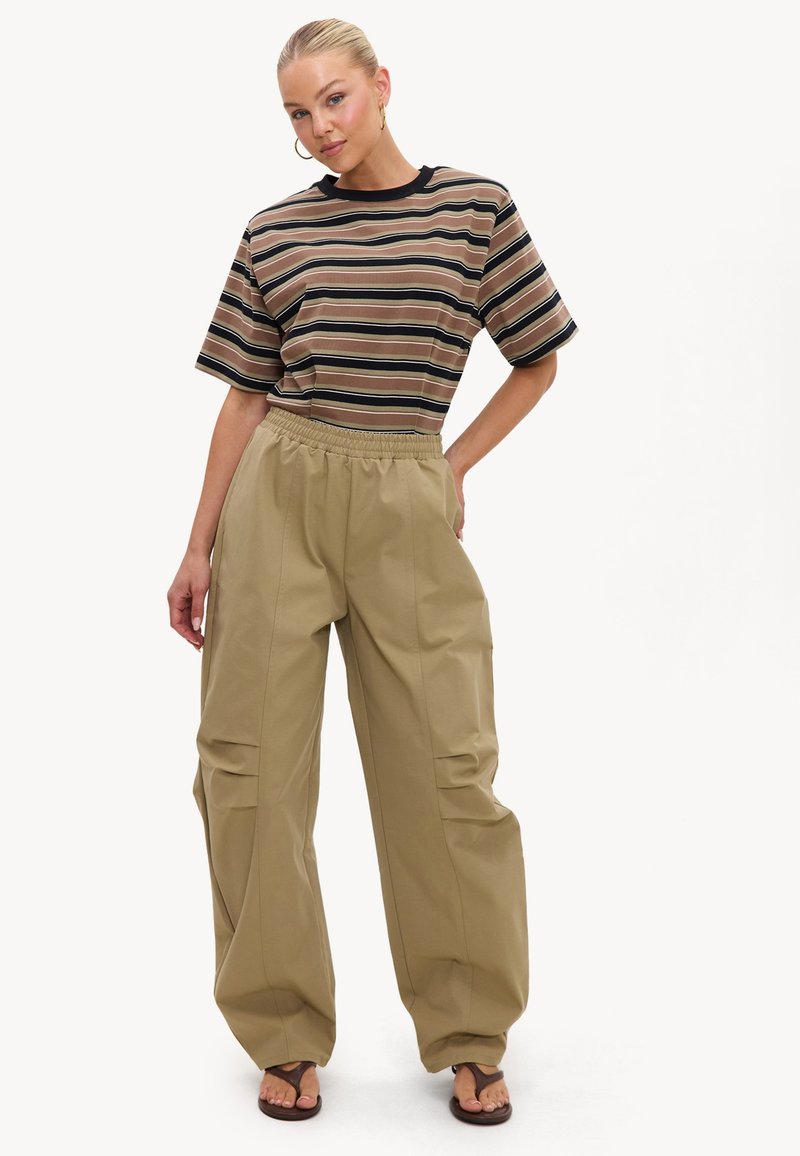 Femme aux cheveux blonds en chignon portant un t-shirt rayé marron, un pantalon beige ample et des sandales foncées, debout avec une main sur la hanche.