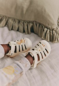 Cream-colored rubber sandals with cutout design and a duck motif. Fastened with a strap, worn with patterned socks.