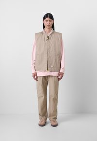 Beige ermeløs vest med glidelås foran og lommer, brukt over en lyserosa langermet skjorte, kombinert med beige bukser og sko.